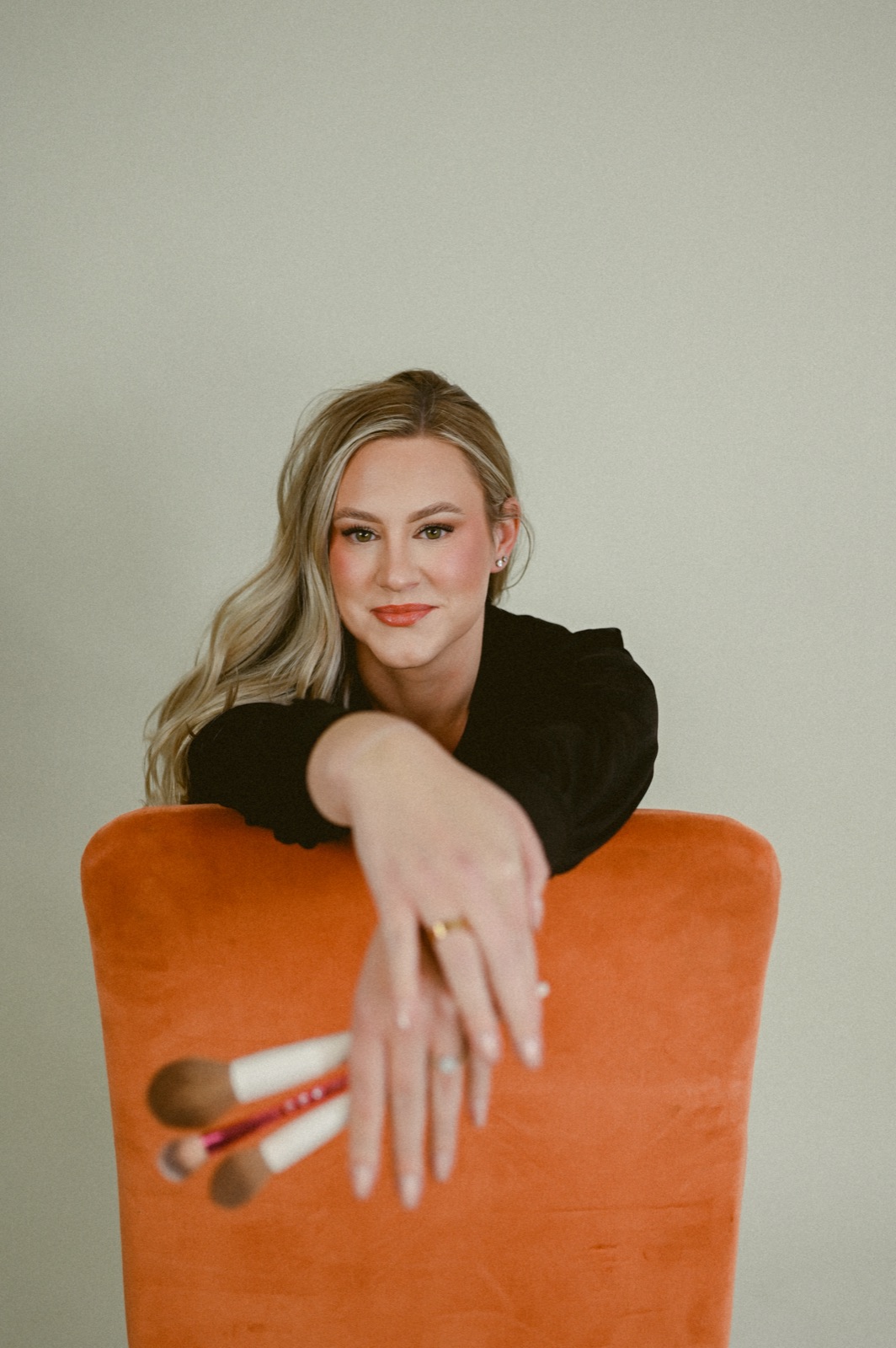 Bailee Newsome smiling, leaning on a chair and holding makeup brushes